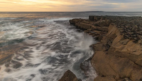 Rocky coastal cliff with waves crashing below during golden hour with distant hills on horizon.