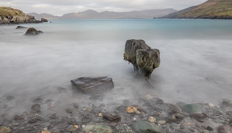 Moss-covered rock jutting from milky seawater with mountains across a bay under overcast sky.