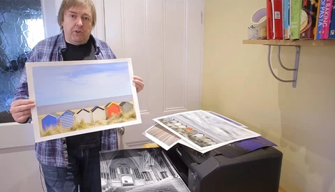 Artist holding a watercolor painting of beach huts against a sky, with additional artwork and prints visible on a desk.