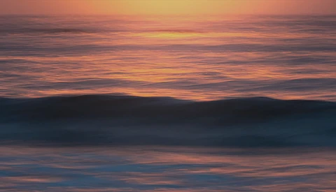Ocean waves rolling toward shore at sunset, with warm orange and pink tones reflecting across the water and sky.