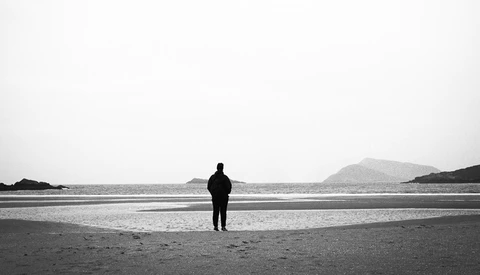 Solitary figure in dark clothing standing on a beach, facing calm water with distant hills.