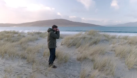 Photographer in dark jacket holding camera to face on sandy beach with dune grass and mountain across bay.