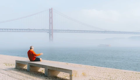 Person in red jacket sitting on concrete bench facing a suspension bridge over water in fog.