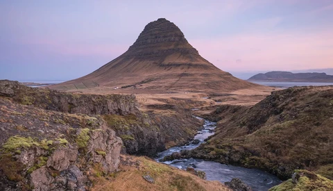 Kirkjufell mountain with snow-dusted slopes rising above a braided river valley at dusk.