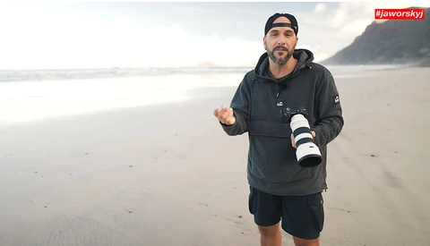 Photographer holding a telephoto lens on a misty beach while gesturing during discussion.