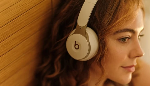 Woman wearing white over-ear headphones, photographed in profile against a warm wooden background.