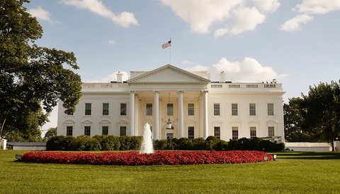 White neoclassical building with columned facade, manicured lawn, and red flowering plants in foreground.
