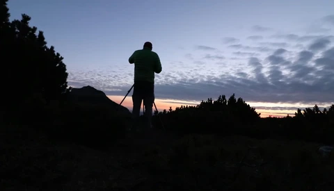 Silhouetted figure standing on elevated terrain with binoculars during twilight, surrounded by forested landscape.