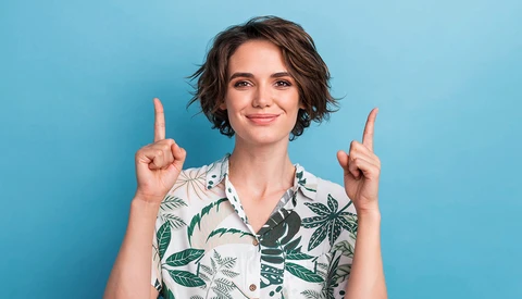 Woman with short dark hair wearing a tropical print shirt pointing upward with both index fingers against a blue background.