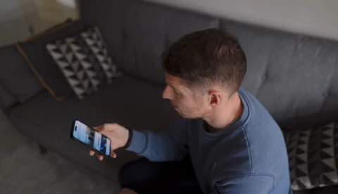 Man seated on a gray sofa looking at a smartphone screen.