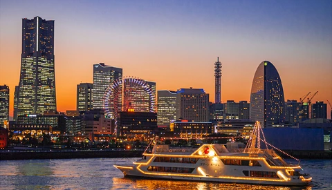 Illuminated ferry boat on water at dusk with city skyline and glowing buildings in background.