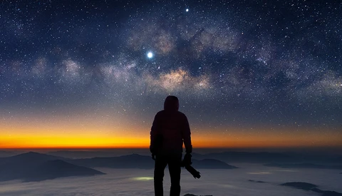 Silhouetted figure standing on a mountain peak facing the Milky Way galaxy at sunrise.