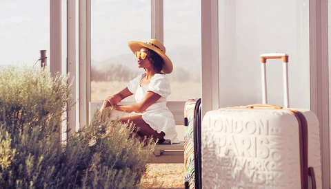 Woman in white dress and straw hat sitting by large windows with lavender plants and luggage beside her.