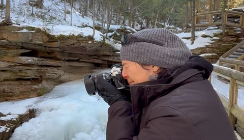 Photographer holding a camera to their face while standing in a snowy forest landscape.