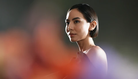 Woman with dark hair wearing white earbuds, looking over shoulder against blurred colorful background.