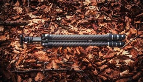 Black telephoto lens with carbon fiber finish and metal bands laid on dried brown leaves.