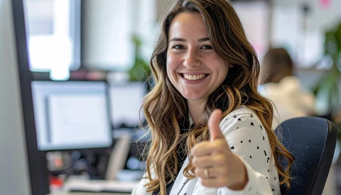 Woman in polka dot blouse giving thumbs up gesture while seated at office desk.