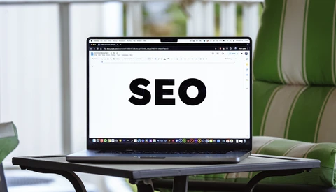 Laptop displaying the word SEO on its screen, positioned on a desk with green striped upholstered furniture visible in the background.
