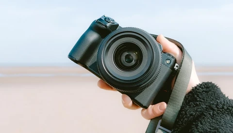 Photographer holding a mirrorless camera with attached lens against a beach backdrop.