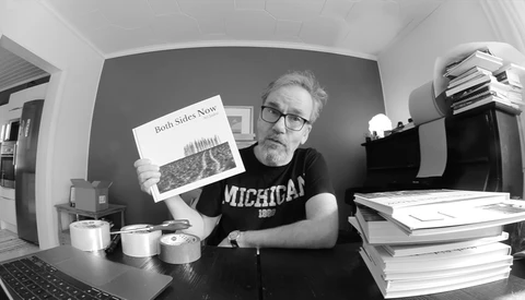 Man in Michigan shirt holding printed publication at desk surrounded by stacks of papers and office supplies.