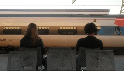Two people seated facing a parked train with tan and blue livery, shot from behind through mesh chairs.
