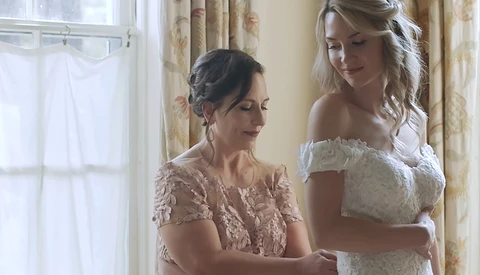 Two women in elegant lace dresses share a moment by a bright window with floral curtains.