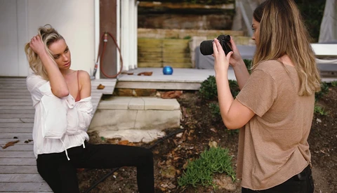 Woman posing outdoors while another woman photographs her with a smartphone.