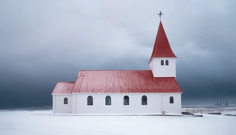White wooden church with red roof and steeple standing alone on a frozen lake under stormy skies.