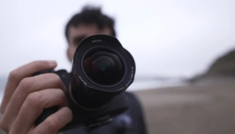Photographer holding a camera with telephoto lens pointed toward the camera in a beach setting.