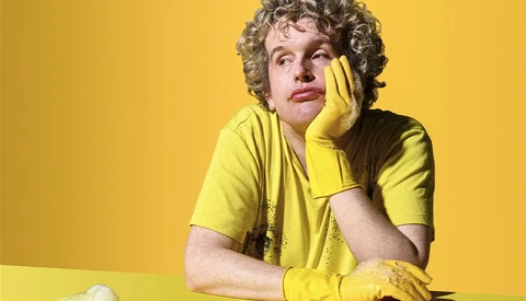 Person in yellow shirt and gloves posing thoughtfully against yellow background.