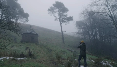 Photographer in yellow beanie shooting in misty hillside landscape with solitary pine tree and stone structure.
