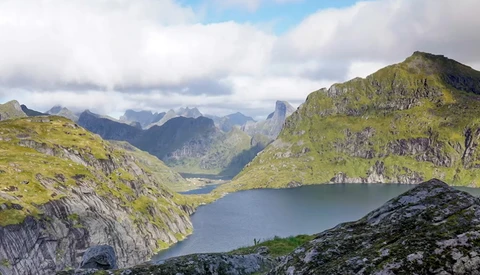 Alpine lake nestled between steep moss-covered mountains under partly cloudy sky.