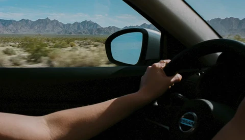 Driver's hand on steering wheel with desert landscape and mountains visible through windshield and side mirror.