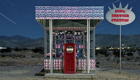 Retro-style service station kiosk in desert landscape at night with neon signage and ornamental patterns.