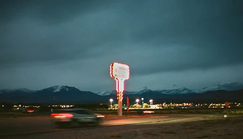Neon-lit roadside sign glowing against dark mountains at dusk with car light trails.