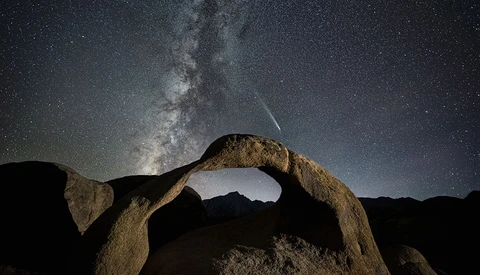 Milky Way galaxy stretching above a natural rock arch formation under a starry night sky.