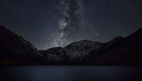 Milky Way galaxy arches over a dark mountain landscape reflected in still water below.