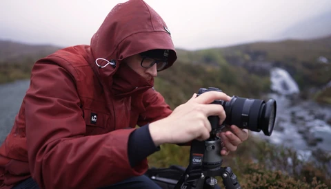 Photographer in maroon jacket and sunglasses operating a mirrorless camera on a tripod in a mountainous landscape with a waterfall.