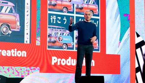 Speaker on stage in front of large red screen displaying colorful vintage cars and production text.