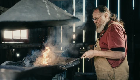 Blacksmith working at an anvil with sparks flying in a workshop lit by overhead lighting.