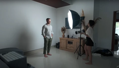 Portrait photography setup with male subject standing in studio with softbox lighting and wooden furniture backdrop.