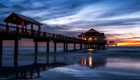 Illuminated pier structures silhouetted against a vibrant sunset sky reflecting in shallow water.