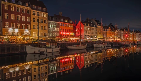 Historic waterfront at dusk with colorful buildings reflected in still water, illuminated by warm lights.