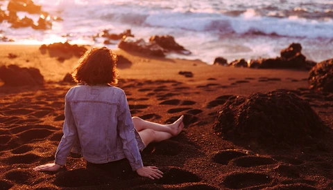 Person sitting on beach sand facing ocean at golden hour sunset.