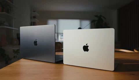 Two MacBook laptops displayed closed on a wooden desk, one silver and one white, photographed in indoor studio lighting.