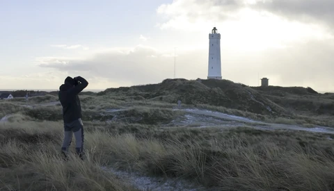 Photographer with camera raised to face standing in coastal dunes with lighthouse on distant hill.