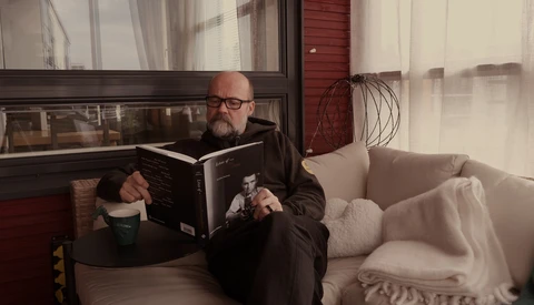 Bearded man wearing glasses reading a book while seated by a window with natural light streaming in.