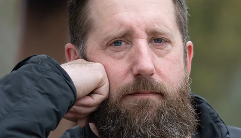 Bearded man with hand raised to face, wearing dark jacket, looking directly at camera with serious expression.