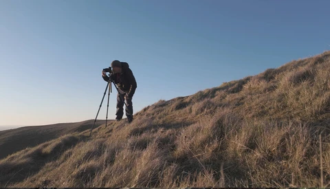 Photographer on a hillside with tripod and camera, capturing the golden-hour landscape in arid terrain.
