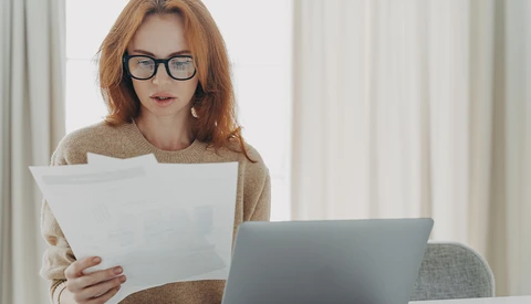Woman with reddish-brown hair and glasses reviewing documents while working at a laptop.